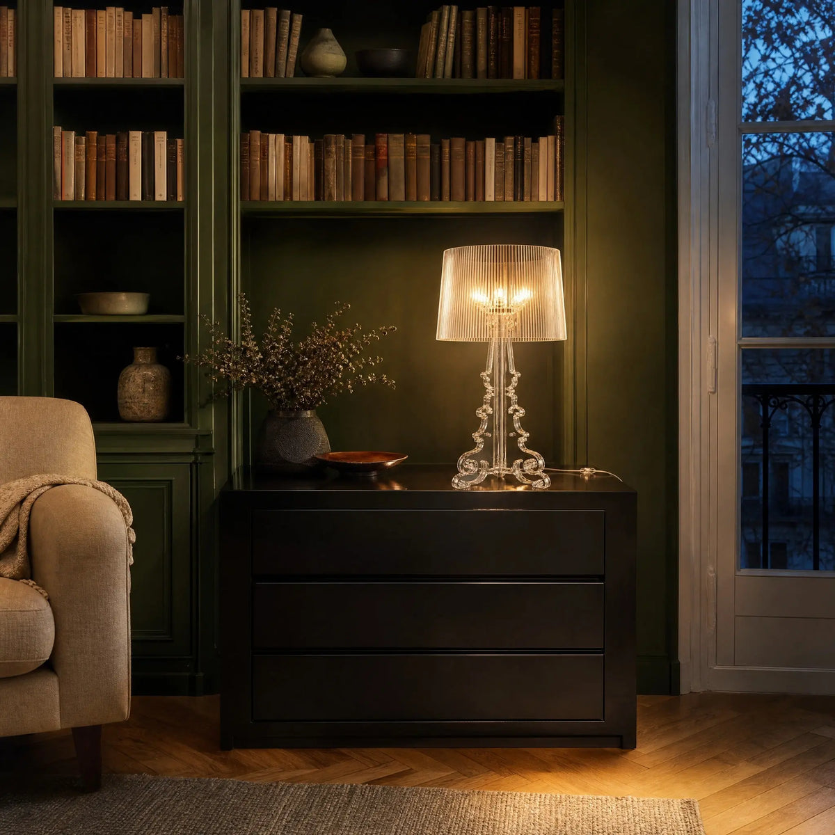 Lampe baroque Rivento transparente allumée sur une commode laquée noire devant une bibliothèque vert anglais