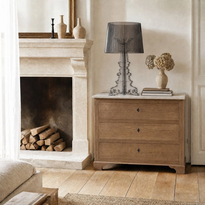 Lampe baroque Rivento en acrylique noir fumé posée sur une commode à plateau de marbre près d'une cheminée en pierre