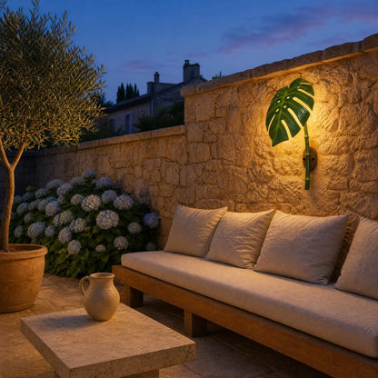 Applique extérieure feuille de Monstera allumée au crépuscule sur un mur de pierre de terrasse, banquette en teck et hortensias | Delvanie